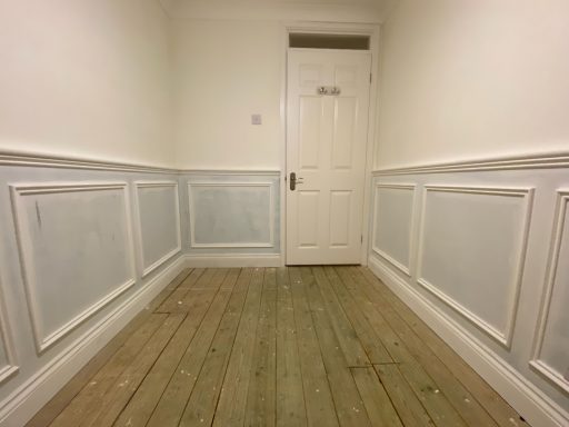 Narrow corridor with wooden flooring and white panelled walls leading to a closed door.
