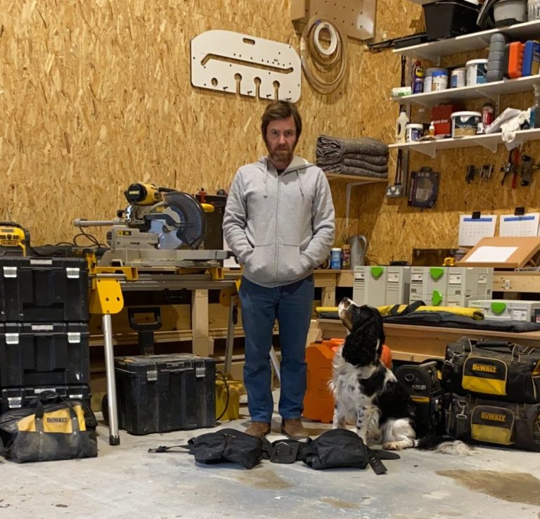 Man standing in a workshop with tools and materials, accompanied by a dog.