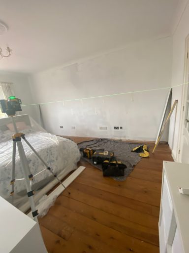 Empty room with a bed, laser on a tripod, and tools on the wooden floor.