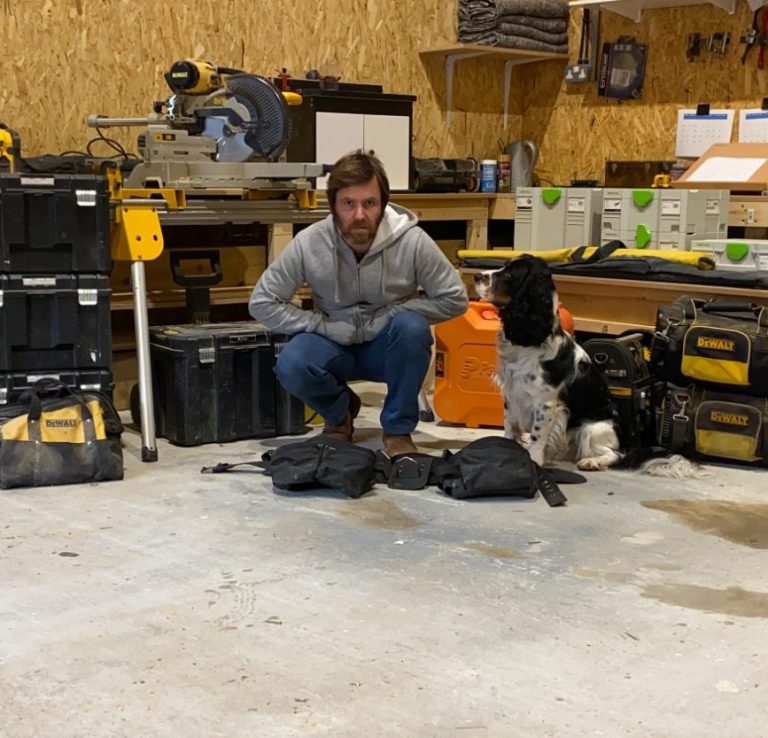 Man crouching beside a dog in a workshop filled with tools and equipment.