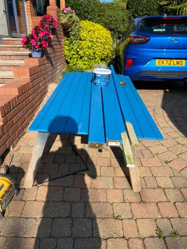 A blue timber with paint cans and tools, surrounded by flowers and a car.
